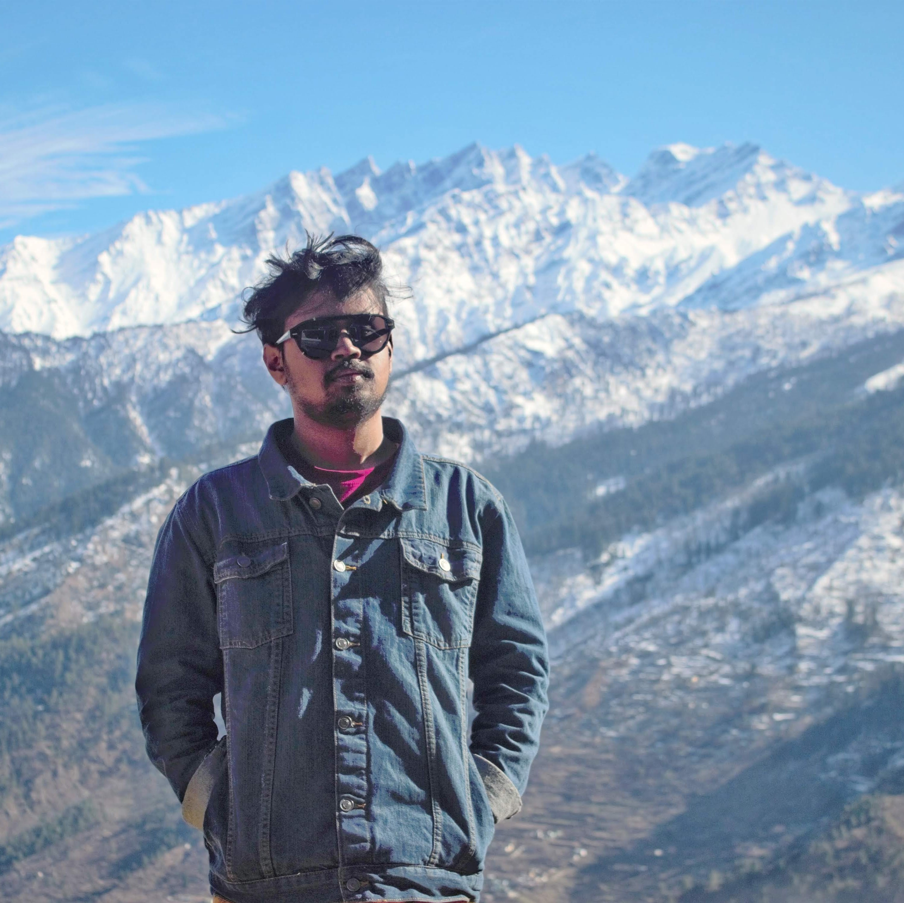 Mobin, a young man, looking at the camrera. He is wearing a dark blue denim jacker, a sunglass, both hands in his pockets. Background shows a snowy mountain of Uttarakhand, India.